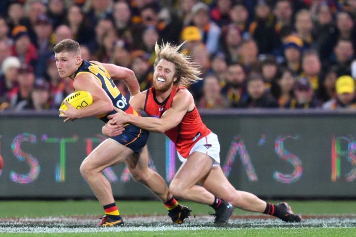 A male AFL player haolds the ball with his left hand as he is tackled around the waist by an opposition player.|700x467