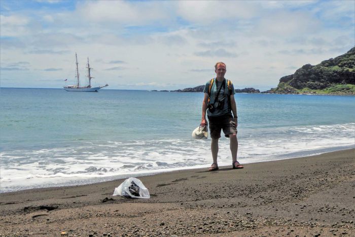 A man in shorts and t-shirt wearing a backpack and camera equipment stands on a black sand beach with a boat in the background|700x467