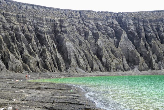 A blue-green crater lake in the foreground with a volcanic rim in the background lined with eroded gullies|700x467