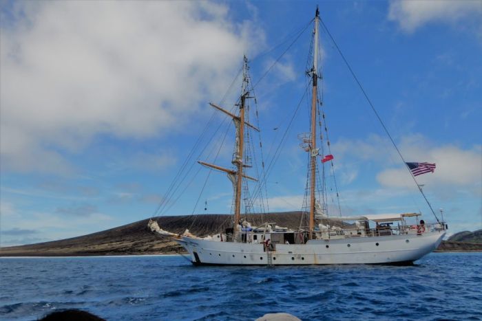 A metal-hulled boat with two masts and a US flag moored next to a low volcanic island|700x467