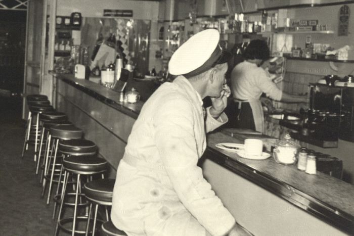 A cafe from 1952, with a milkman sitting at the bench drinking coffee.|700x467